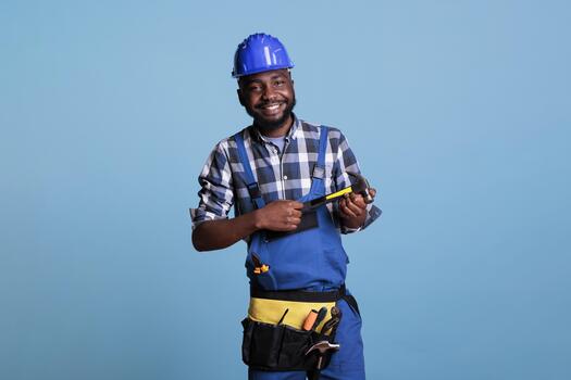 happy construction worker smiling optimistic against blue isolated background studio of african american man wearing hard hat and tool belt holding hammer with both hands photo